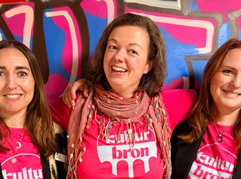 Tre personer står tillsammans framför en färgglad graffitivägg, alla bär rosa t-shirts med texten 'kultur bron'.