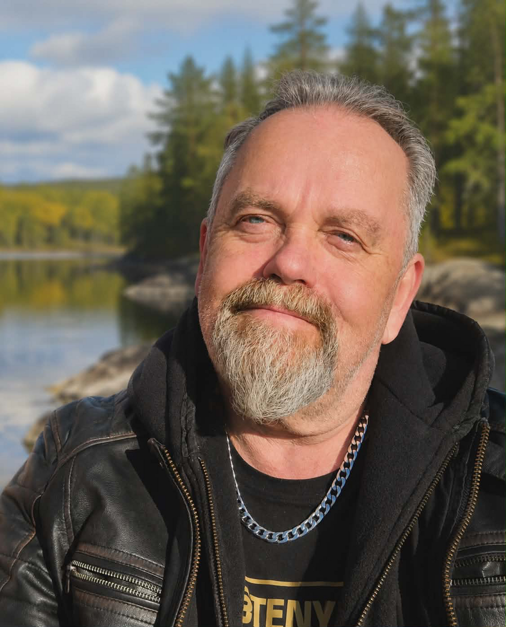 Man med grått skägg och läderjacka poserar utomhus vid en sjö med skog i bakgrunden.