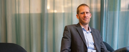 Man i kostym sitter avslappnat i en fåtölj framför ett blågrönt draperi.