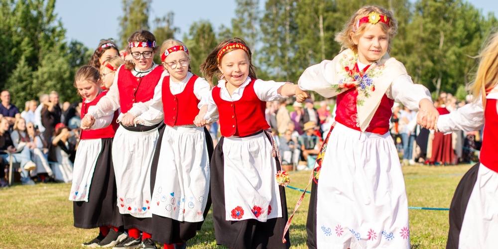 Barn i traditionella folkdräkter dansar i ring utomhus på en gräsmatta inför publik.