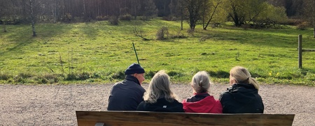 Fyra personer sitter på en bänk och tittar ut över en grön äng under en molnig himmel.