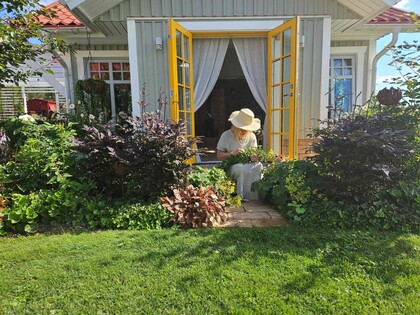 En person med solhatt sitter vid ett bord utanför ett litet hus med öppna gula dörrar, omgiven av grönska.