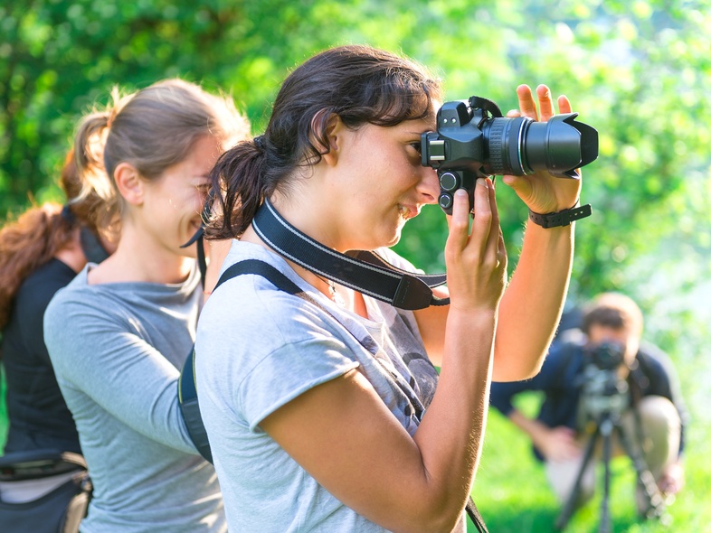 Två kvinnor fotograferar utomhus i grönskande miljö, med en tredje person i bakgrunden.