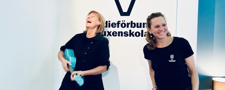 Två kvinnor står framför en vägg med logotypen 'SV', en spelar ukulele och båda ser glada ut.