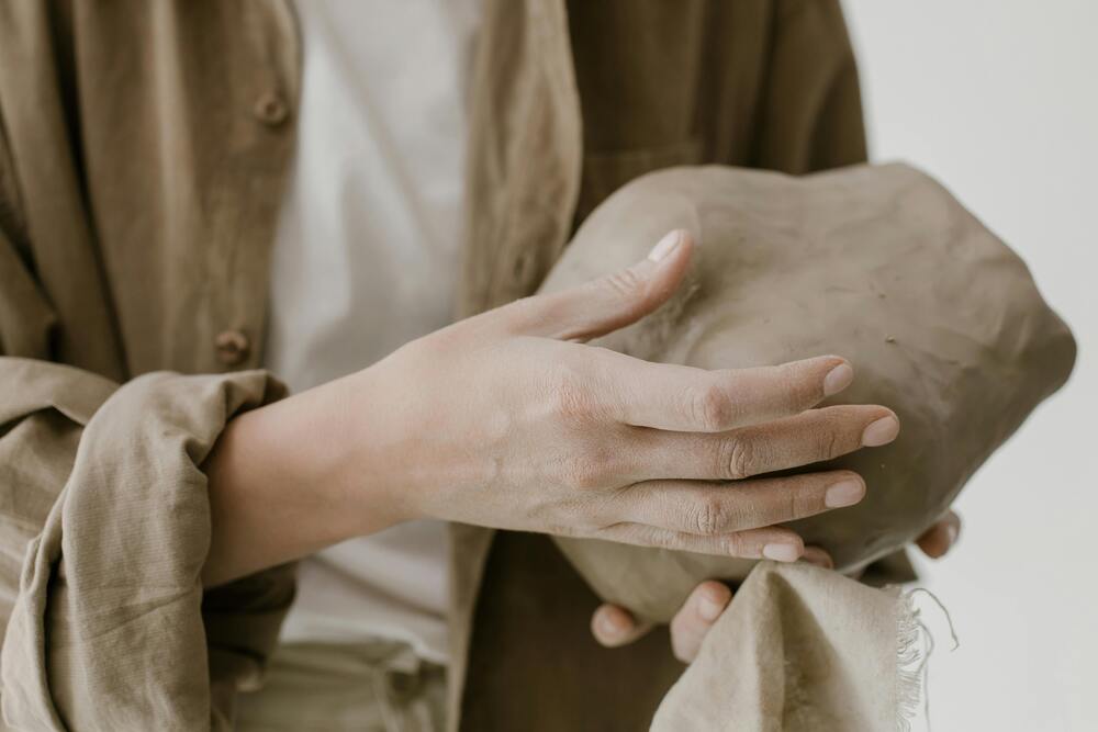 En person håller en stor klump lera i händerna, klädd i beige och vitt.