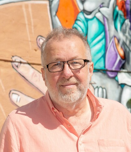 Man med glasögon och skägg i ljusrosa skjorta står framför en färgglad graffitivägg utomhus.