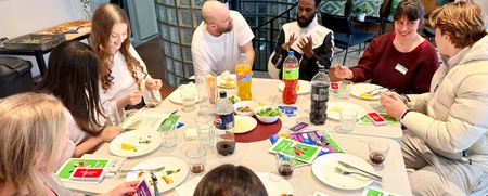 En grupp unga vuxna sitter runt ett bord och spelar ett kortspel, med mat och dryck på bordet.