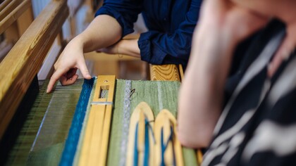 Två personer arbetar vid en vävstol, en pekar på tyget som vävs i gröna och blå nyanser.