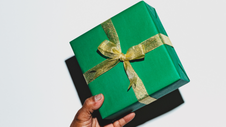 En hand håller ett grönt inslaget paket med ett guldigt band mot vit bakgrund.