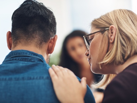 En kvinna tröstar en man genom att lägga handen på hans axel i en stödjande miljö.