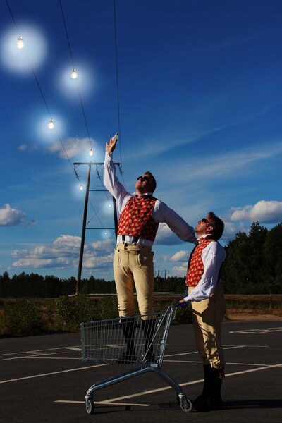 Två personer i färgglada västar står i en kundvagn på en tom parkering och sträcker sig mot glödlampor i himlen.