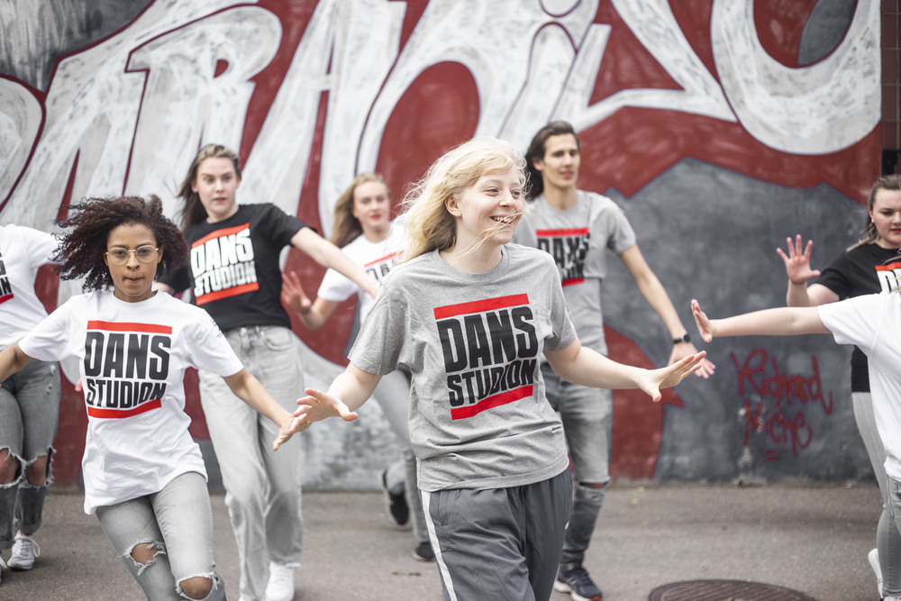 En grupp unga personer dansar energiskt utomhus framför en graffitivägg, alla bär t-shirts med texten 'DANS STUDIO'.