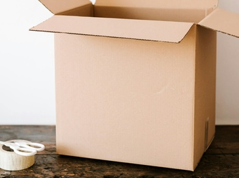 En öppen kartong står på ett träbord bredvid en sax och en rulle tejp.