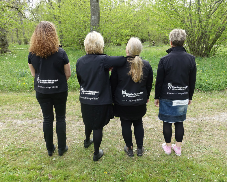 Fyra kvinnor står utomhus med ryggen mot kameran, iförda svarta jackor med text på ryggen.