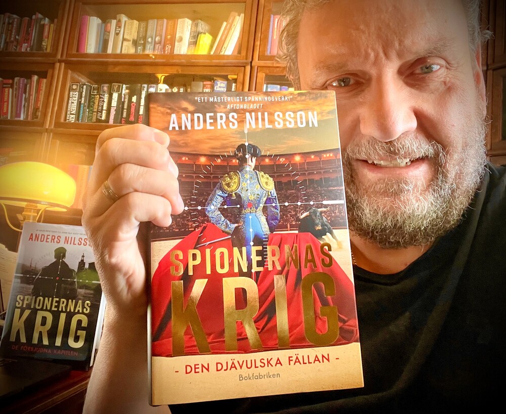 En person håller upp boken 'Spionernas krig' av Anders Nilsson i ett rum med bokhyllor och böcker.