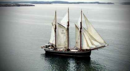 Segelfartyg med tre master seglar på lugnt vatten med öar i bakgrunden.