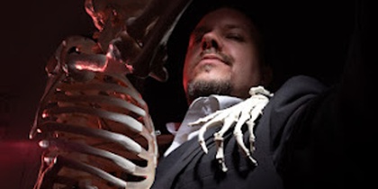 En person i mörk kostym håller ett skelett, med skelettets hand på personens axel. Ansiktet är suddat.