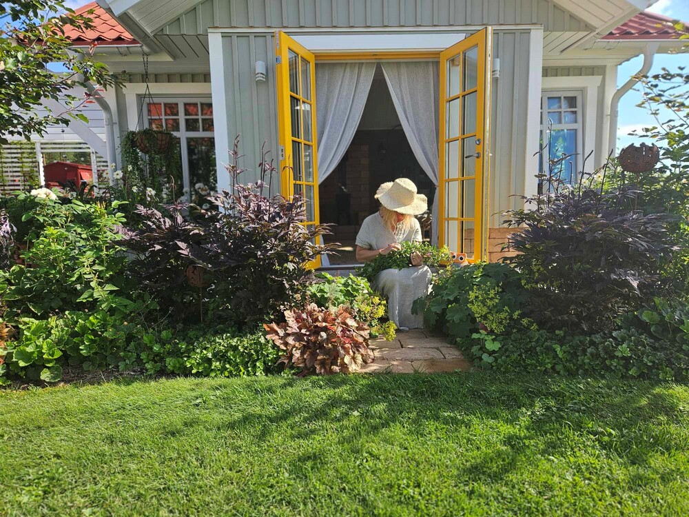 En person med solhatt sitter vid ett bord utanför ett litet hus med öppna gula dörrar, omgiven av grönska.