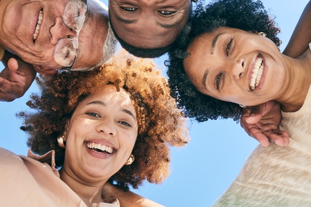 Fyra personer står i en cirkel och tittar ner mot kameran, alla ler mot varandra under en klarblå himmel.