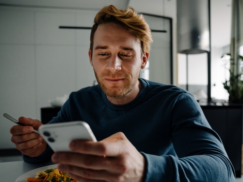 Man sitter vid ett bord, äter mat och tittar på sin mobiltelefon i ett modernt hem.