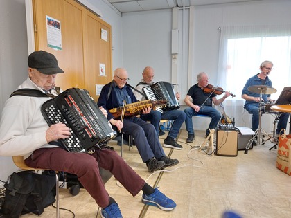 Fem personer spelar olika instrument i ett ljust rum, inklusive dragspel, nyckelharpa, fiol och trummor.