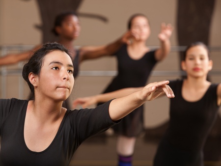 Fyra unga personer i svarta danskläder övar balett i en dansstudio, fokus på en person i förgrunden.