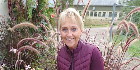 Person sitter på en blomlåda utomhus omgiven av prydnadsgräs och blommor.