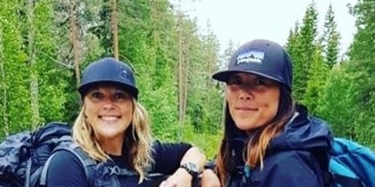Två kvinnor med ryggsäckar står på en träbro i skogen vid en forsande å.
