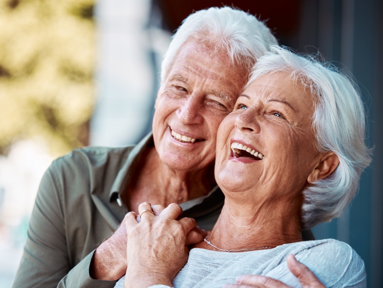 Ett äldre par skrattar och kramas utomhus i dagsljus.