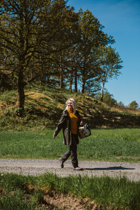 Kvinna promenerar på en grusväg i naturen under en solig dag, omgiven av grönt gräs och träd.