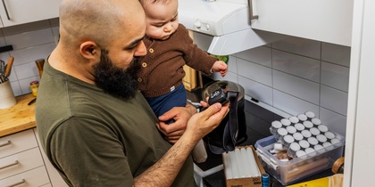 Vuxen person med skägg, bandage runt armbågen och olivgrön tröja håller i ett litet barn i brun stickad kofta. Den vuxne betraktar en pannlampa. Miljön är ett kök med stearinljus, värmeljus på köksbänk bredvid spis.