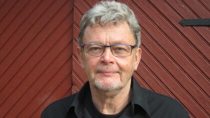 Person med grått hår och svart skjorta står framför en röd trävägg med diagonala brädor.