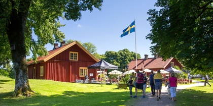 Folk promenerar mot röda trähus med svensk flagga, soligt väder och grönskande omgivning.