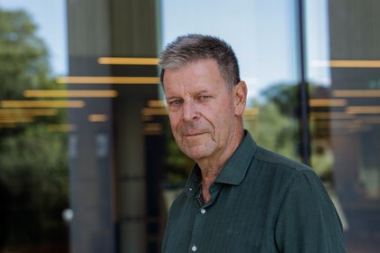 En man i grön skjorta står utomhus framför en glasvägg med reflektioner och träd i bakgrunden.