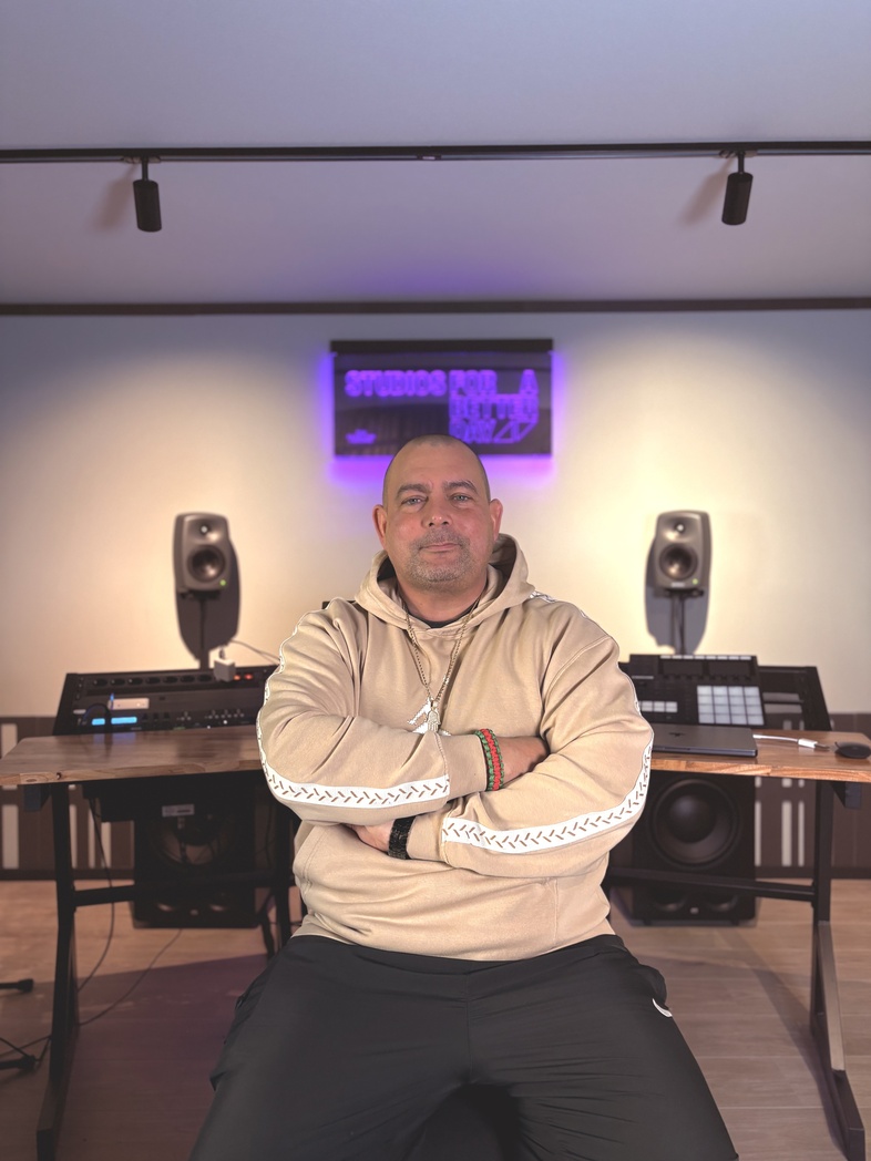 Man i beige hoodie sitter med armarna i kors i en musikstudio med högtalare och utrustning bakom sig.