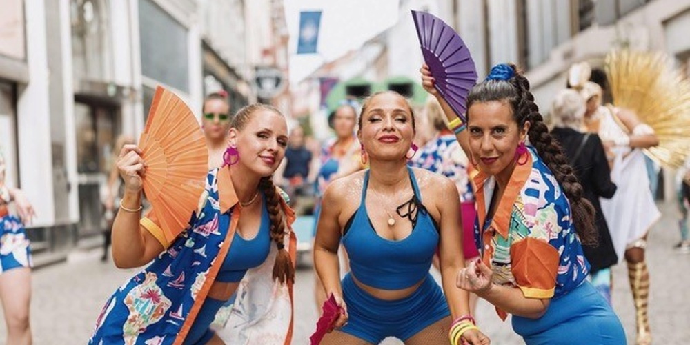 Tre kvinnor i färgglada kläder dansar med solfjädrar på en stadsgata under en festival.