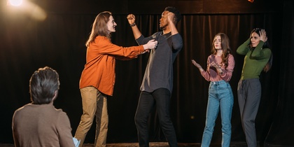 Fyra personer står på en scen och spelar teater, medan en person sitter och tittar på dem.