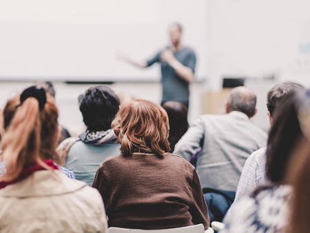 En grupp människor sitter och lyssnar på en person som håller en presentation i ett ljust rum.
