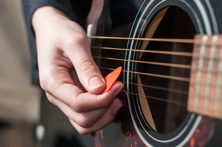 En hand spelar på en akustisk gitarr med ett orange plektrum.