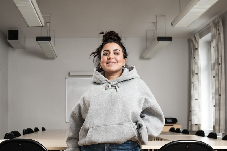 Ung kvinna i grå huvtröja står leende i ett tomt klassrum med bord och stolar.