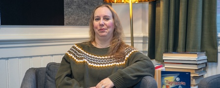 En kvinna med axellångt blont hår sitter i fåtölj med. Bakom henne syns en lampa och en trave böcker.