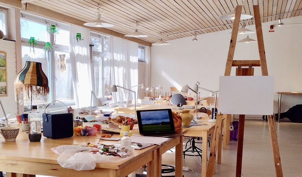 Ljust konstnärsateljé med arbetsbord, staffli, målarfärger och dator på borden.