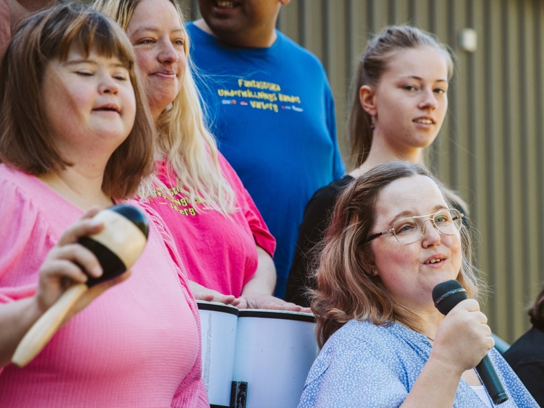 En grupp människor, flera med mikrofon och musikinstrument, står utomhus och sjunger tillsammans.