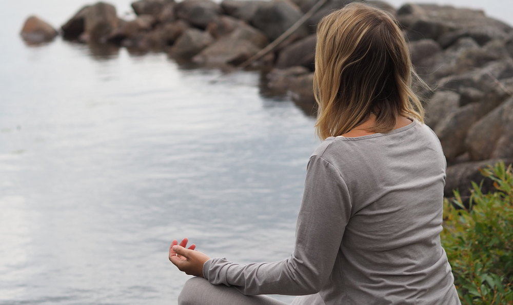 Person sitter i meditationsställning vid en sjö med stenar i bakgrunden.