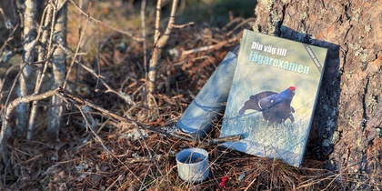 En bok med en fågel på omslaget, en termos och en kopp står lutade mot ett träd i skogen.