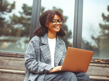 Leende person med glasögon sitter utomhus på trappa och arbetar på en bärbar dator.