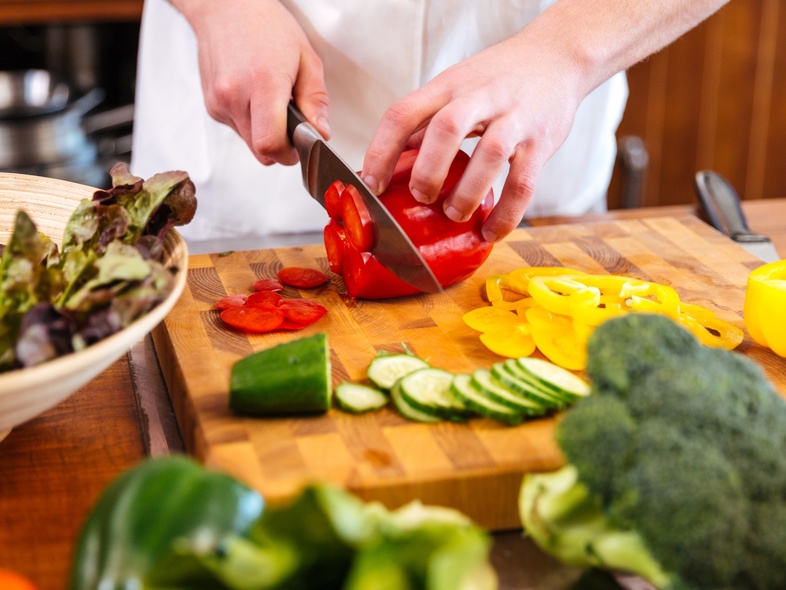 En person skär röda paprikor på en skärbräda omgiven av olika grönsaker i ett kök.