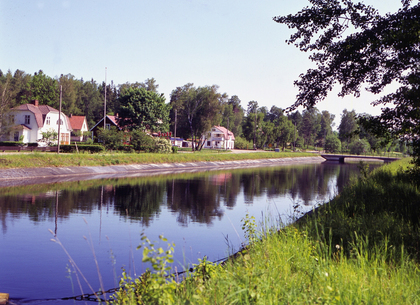 En lugn kanal med speglingar, grönt gräs och hus i bakgrunden under en klar himmel.