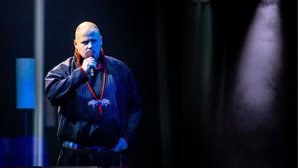 Person står på scen och talar i mikrofon under stark belysning.