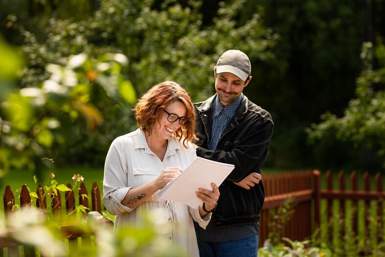 Två personer står utomhus vid ett staket, en kvinna skriver på en surfplatta medan en man tittar på.